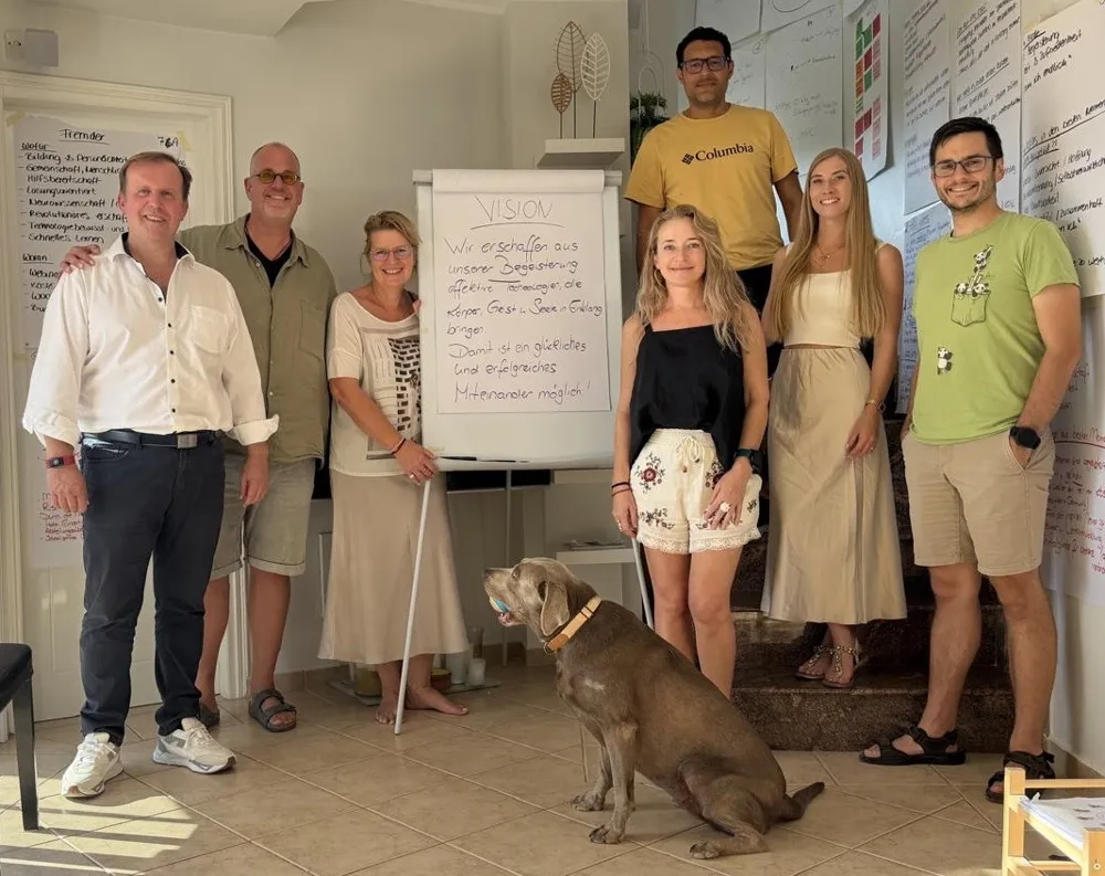 Das Team von KOSYS® Ltd. mit einem Hund vor einem Flipchart. Darauf steht: Vision – Wir erschaffen aus unserer Begeisterung effektive Technologien, die Körper, Geist & Seele in Einklang bringen. Damit ist ein glückliches und erfolgreiches Miteinander möglich!